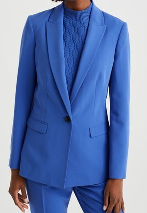 Vrouw draagt een felblauwe, op maat gemaakte blazer over een getextureerde gebreide trui, met bijpassende blauwe broek tegen een effen achtergrond.
