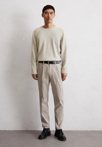 Licht beige, textuurgegeven, lang-mouw overhemd, crème tapered broek, zwarte riem en zwarte veterschoenen op een effen achtergrond.