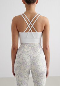 Witte sport-BH met gekruiste rugbanden, gecombineerd met strakke legging met een pastelkleurig bloemenpatroon op een lichte achtergrond.