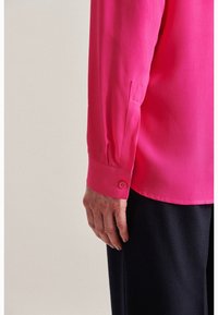 Camisetta a maniche lunghe di un brillante colore rosa, realizzata in tessuto morbido, con dettagli sui polsini abbottonati e una vestibilità comoda, abbinata a pantaloni scuri.