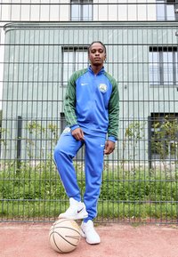 Nike tracksuit in blue and green fabric, featuring a zip-up jacket, logo detail, and matching pants. White sneakers and a basketball on the ground.