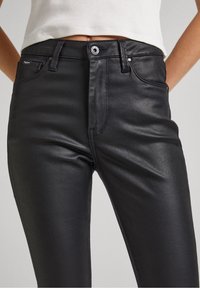 Mujer vistiendo pantalones negros de talle alto con acabado brillante, con botón frontal, cremallera, trabillas para cinturón y bolsillos frontales, combinados con un top blanco corto.