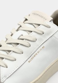 Sneaker in pelle bianca con lacci beige, logo in rilievo sulla linguetta e dettaglio dorato con la data sul lato. Texture liscia con design minimalista.