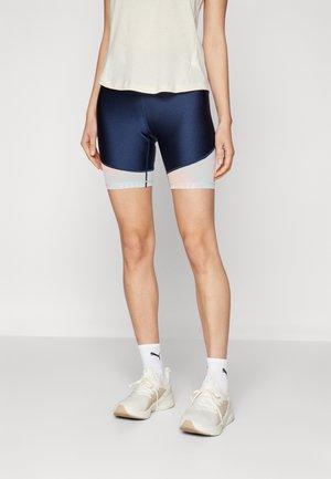 Personne portant un cuissard de cyclisme bleu marine et gris clair, un haut blanc sans manches, des chaussettes blanches Puma et des chaussures de sport blanches, debout sur un fond uni.