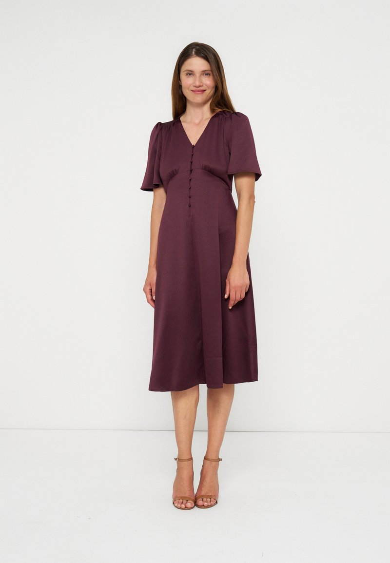 Femme debout portant une robe prune mi-longue avec manches courtes, boutons sur le devant, et des talons à bride à la cheville couleur beige sur un fond blanc uni.