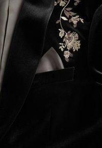 Black velvet blazer featuring floral embroidery in gold and cream tones on the lapel, with a contrasting satin pocket square.