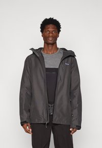 Čierna nepremokavá bunda s nastaviteľnou kapucňou, zipsovými vreckami a elastickými manžetami. Nosí sa cez šedý top, určená na vonkajšie nosenie.