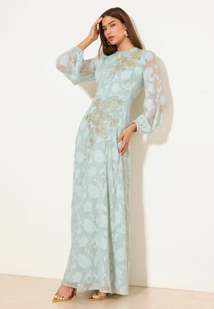 Frau präsentiert ein hellblaues, florales, transparentes Maxikleid mit langen Ärmeln, goldener Stickerei und goldenen Sandalen mit offener Spitze vor einer weißen Wand.