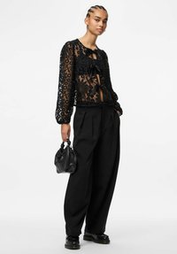 Svart bluse i blonder med blomstermønster, lange ermer og knyting foran, kombinert med løstsittende svarte bukser og en liten svart håndveske.