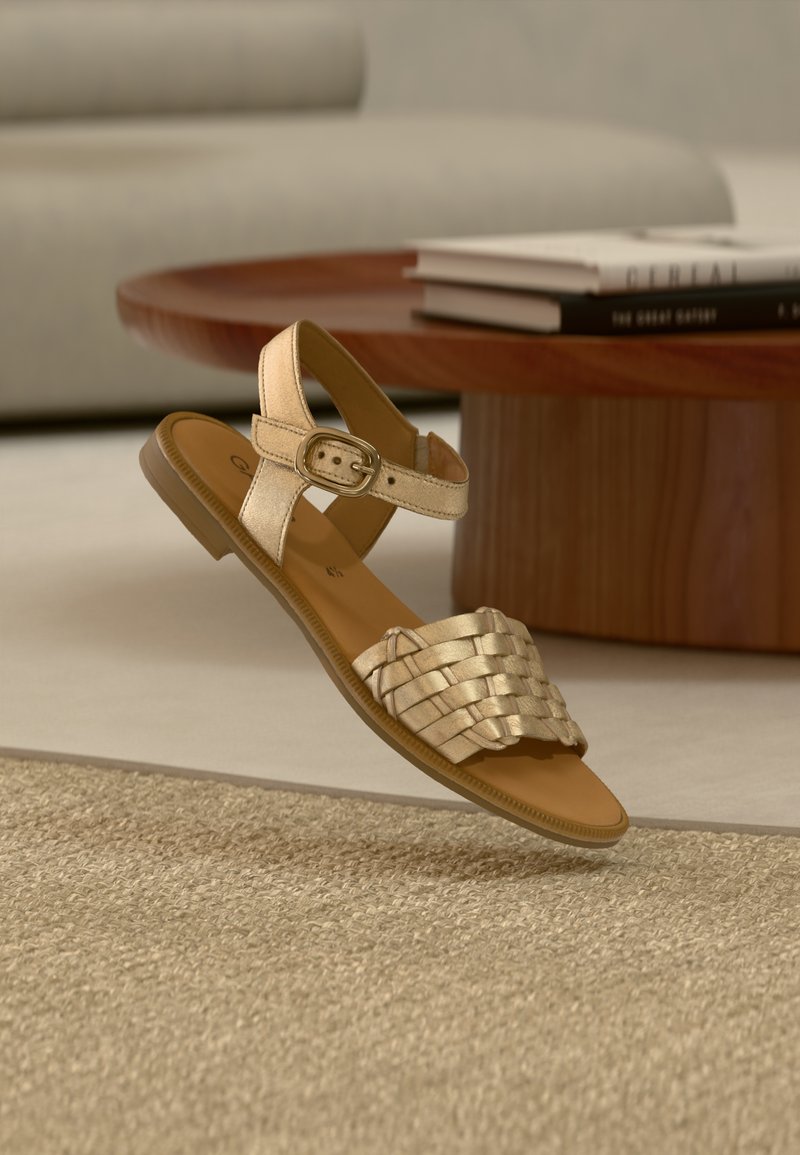Χρυσό μεταλλικό επίπεδο σανδάλι με πλεκτό σχέδιο λουριών, ρυθμιζόμενο πόρπη στον αστράγαλο και μαλακό ταμπά πέλμα σε υφασμένο χαλί.