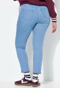 Ljusblå skinny jeans med hög midja, prydda med en rad dekorativa nitar på sidan, kombinerade med bruna och vita sneakers.