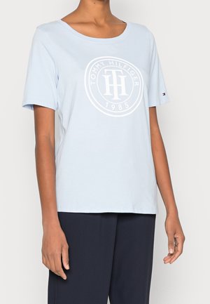 Personne portant un t-shirt Tommy Hilfiger bleu clair avec un logo et un texte blancs, associé à un pantalon bleu marine foncé, debout devant un fond uni.