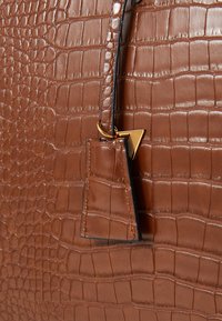 Bruine tas van krokodillenembossed leer met een kleine bijpassende leren label en een gouden driehoekige charme die met een ring is bevestigd.