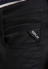 Jeans en denim noir avec une petite poche arborant une étiquette blanche "REPLAY". Le tissu a une texture lisse avec des détails de couture minimaux.