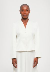Frau mit rasiertem Kopf, die ein maßgeschneidertes weißes Blazer mit Knöpfen und einen passenden Rock trägt, steht vor einem schlichten weißen Hintergrund.