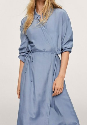 Frau trägt ein hellblaues Wickelkleid mit langen Ärmeln, einem Kragen, das in der Taille gebunden ist und lockere, hochgekrempelte Ärmel hat.