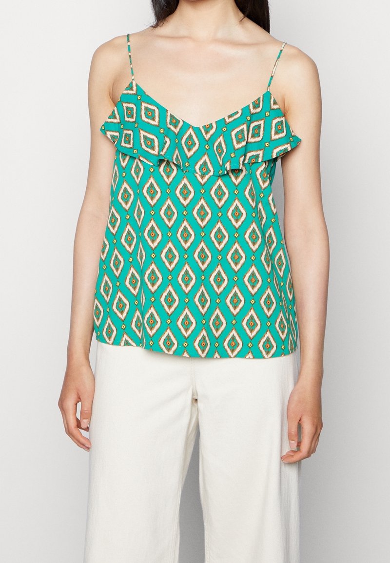 Femme portant un haut vert sans manches avec des motifs géométriques en forme de losanges et un détail volanté, associé à un pantalon blanc ample.