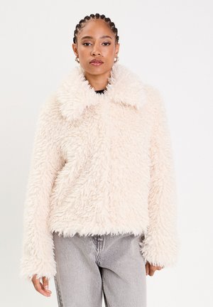 Veste en fausse fourrure de couleur rose clair avec un extérieur texturé et duveteux, un large col et des manches longues. Associée à un pantalon en jean gris.