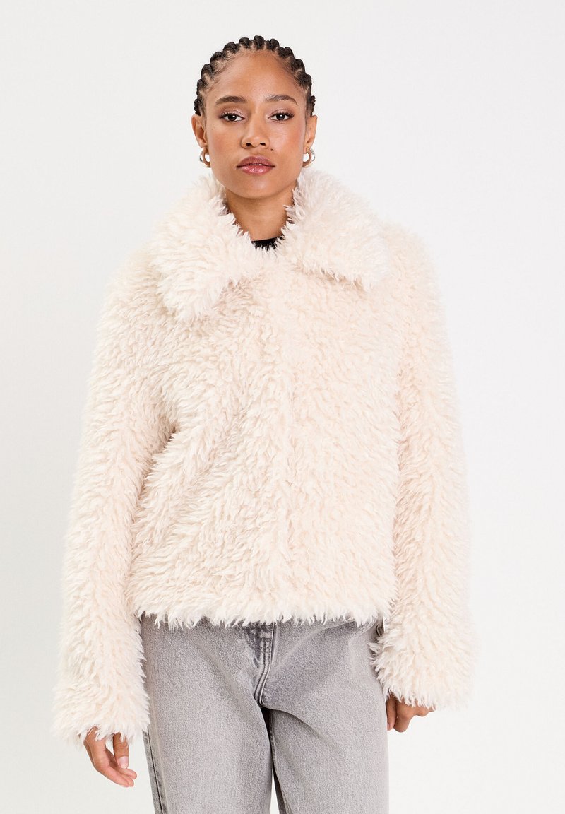 Veste en fausse fourrure de couleur rose clair avec un extérieur texturé et duveteux, un large col et des manches longues. Associée à un pantalon en jean gris.