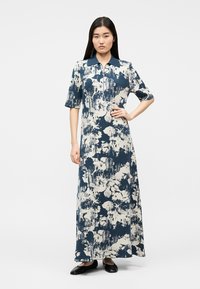 Woman standing with hand on hip wearing a blue and white abstract print long dress with short sleeves and black flats.
