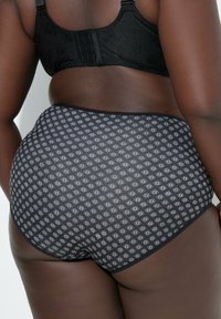 Mujer con ropa interior negra de cintura alta con estampado y un sujetador negro, vista desde atrás sobre un fondo liso.