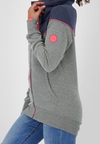 Felpa con zip grigia e blu navy con dettagli rosa, colletto alto e texture morbida. Presenta una toppa rotonda con logo sulla manica e tasche laterali.
