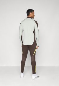 Tenue de sport composée d'un haut à manches longues gris clair et d'un pantalon marron ajusté avec des accents jaunes et un design en panneaux.