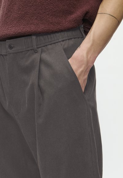 O mână se odihnește în buzunarul din față al unui pantalon gri închis, purtat cu un top texturat de culoare vișinie și un tatuaj subțire vizibil pe braț.