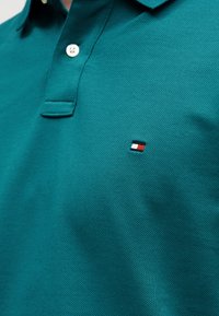 Μπλουζάκι πόλο από βαμβάκι σε τιρκουάζ με υφή, με κούμπωμα δύο κουμπιών και μικρό κεντητό λογότυπο σε κόκκινο, λευκό και μαύρο.