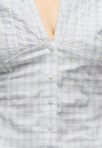 Λευκό πουκάμισο με κουμπιά και ανοιχτό γαλάζιο καρό μοτίβο, φορεμένο με το επάνω κουμπί ανοιχτό, αποκαλύπτοντας μικρή περιοχή στο στήθος.
