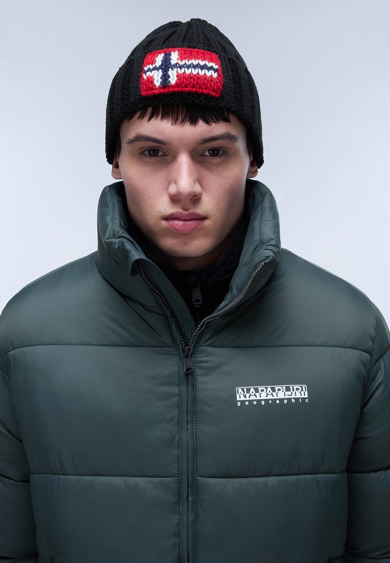 Chaqueta acolchada verde con cuello alto y cierre de cremallera. Gorro de lana negro con un patrón de bandera noruega en rojo y blanco.