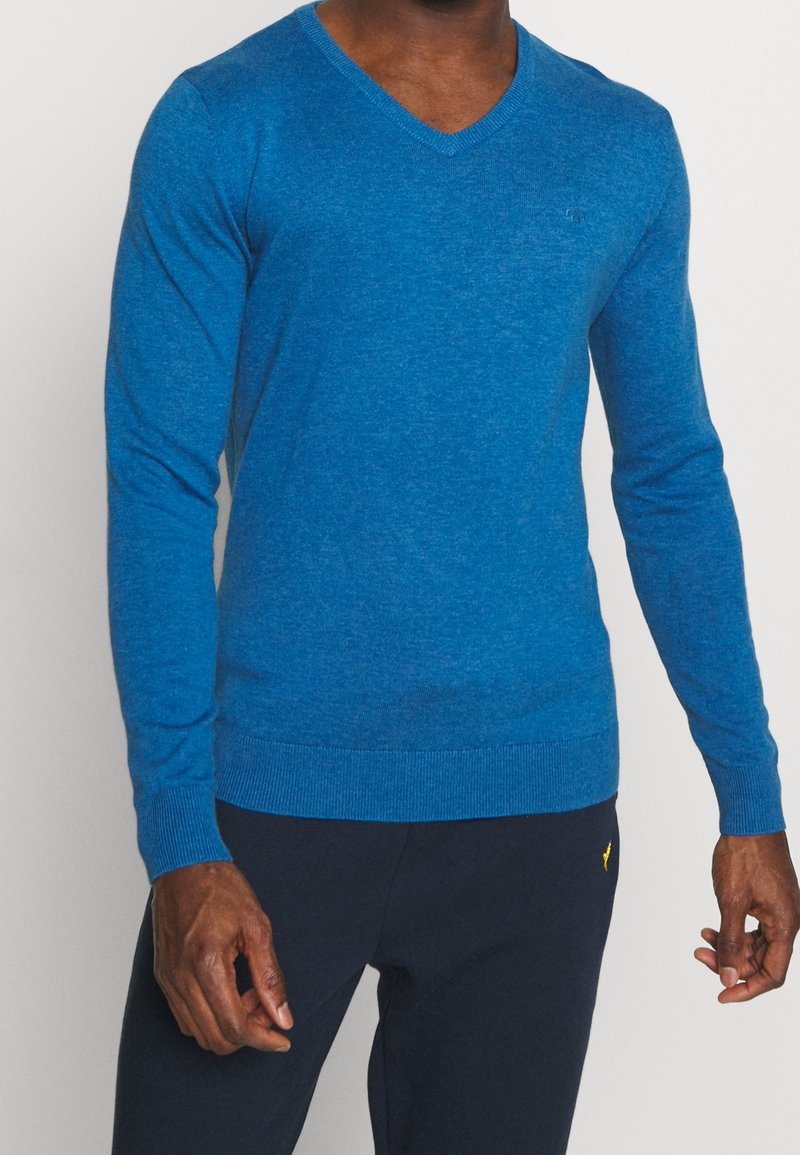 Mann trägt einen enganliegenden blauen V-Ausschnitt-Pullover und dunkelblaue Hose, steht vor einem schlichten Hintergrund.