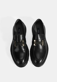 Schwarze Lederloafers mit einem goldfarbenen Hardware-Akzent, runder Zehenform und sichtbaren Nähten an der Sohle. Branding innen sichtbar.