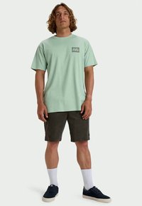 Mann steht mit einem hellgrünen kurzärmeligen Hemd, dunklen Shorts, weißen Socken und schwarzen Turnschuhen vor einem einfarbigen hellen Hintergrund.