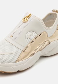 Hvit sneaker med teksturert mesh-overdel, gullfiner, glidelås for lukking og gummisåle med sirkulære mønstre.