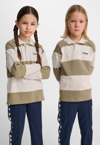 Randiga långärmade polo skjortor i beige och olivgrön, kombinerade med navybyxor som har en vit chevrondesign längs sidorna.