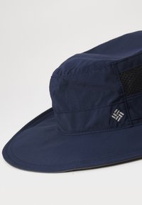Marineblå bøttehatt laget av lett stoff, med en pustende mesh-bånd og brodert logo-detalj på siden.