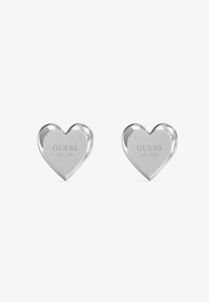 Orecchini a forma di cuore in argento con superficie lucida e incisione del testo "GUESS EST. 1984" sulla parte anteriore.