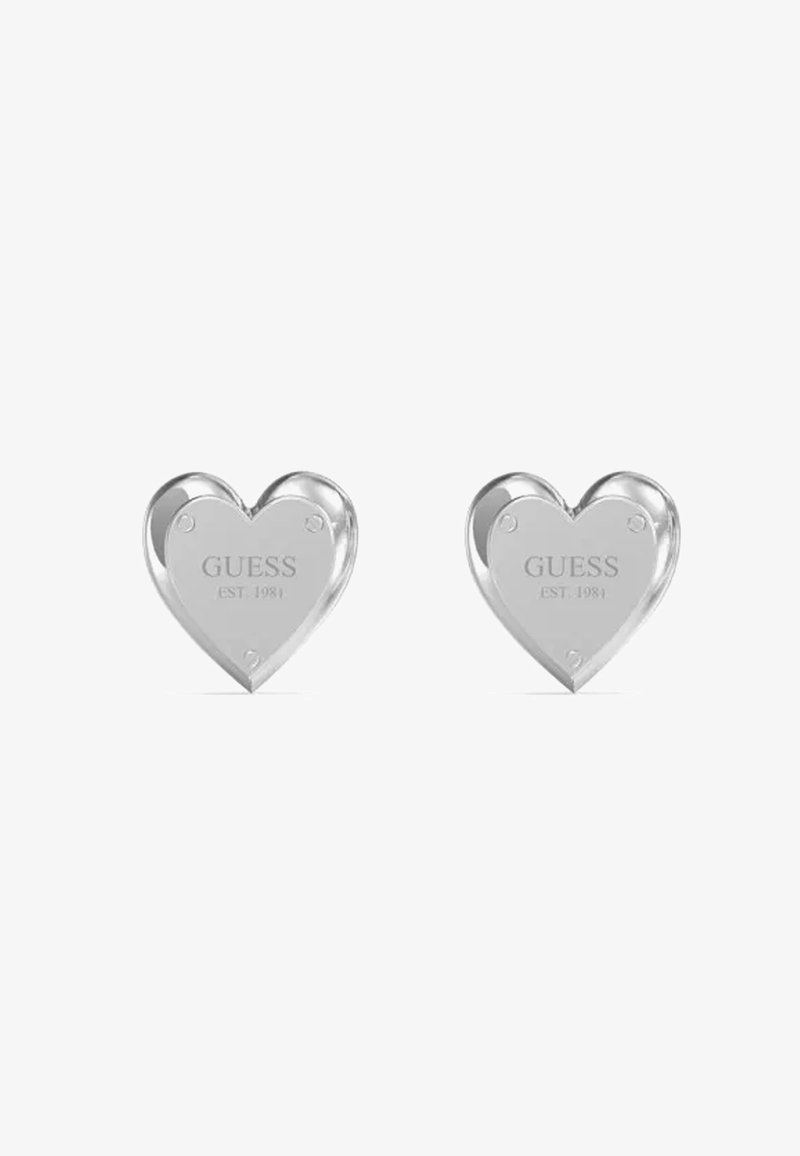 Orecchini a forma di cuore in argento con superficie lucida e incisione del testo "GUESS EST. 1984" sulla parte anteriore.