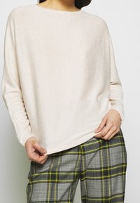 Maglietta a maniche lunghe di un beige chiaro con una consistenza morbida, scollatura rotonda e vestibilità rilassata, abbinata a pantaloni a quadri grigi con dettagli verdi.