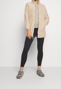 Beżowa polarowa kurtka z kapturem, z przodu na zamek błyskawiczny i bocznymi kieszeniami, w połączeniu z czarnymi legginsami i szarymi butami sportowymi.