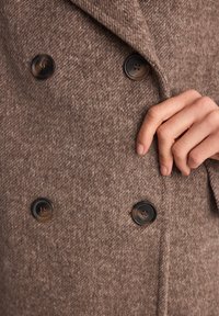 Brązowy płaszcz z wełny z dodatkiem innych włókien, charakteryzujący się teksturowanym wykończeniem, podwójnym zapięciem na guziki i kontrastującymi czarnymi guzikami. Ręka umieszczona na tkaninie.