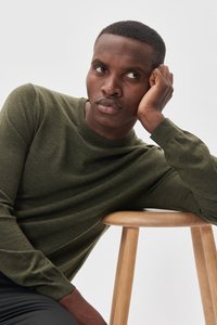 Mörkgrön stickad tröja med rund halsringning, med en slät textur och åtsittande ärmar. Trästool i förgrunden.