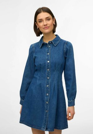 Mujer con vestido de mezclilla azul de manga larga, abotonado y con cuello, posando frente a un fondo blanco liso.