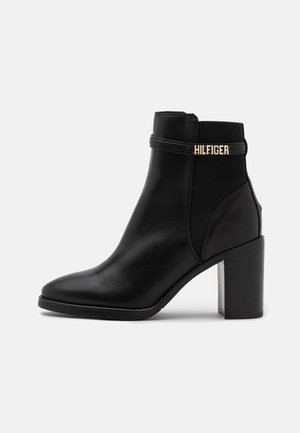 Zwarte leren enkellaars met een puntige neus, blokhak, elastische zijpanelen en een gouden "HILFIGER" logo-accent. Glad oppervlak.