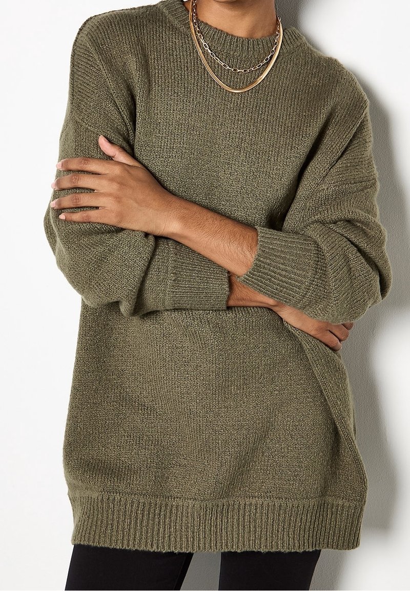 Personne portant un pull en maille vert olive avec poignets et ourlet côtelés, posant les bras croisés et portant des colliers en or superposés.