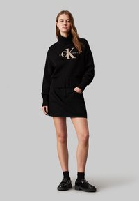 Czarny sweter z dzianiny z prążkowanym wykończeniem, z beżowym logo Calvin Klein. W zestawie czarna spódnica i czarne masywne buty.