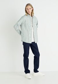 Striped button-up shirt in green and white, made of lightweight fabric; paired with dark jeans and white sneakers.