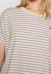 Pasiasta koszulka w kolorze beżowym, białym i czarnym; krótki rękaw; okrągły dekolt; małe haftowane logo na piersiach; miękka, casualowa faktura materiału.