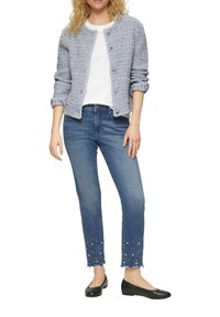 Helles bläuliches, strukturiertes Jacket mit rundem Ausschnitt und Knöpfen, weißes T-Shirt, dunkelblaue Jeans mit Perlenakzenten am Saum und schwarze Ballerinas.